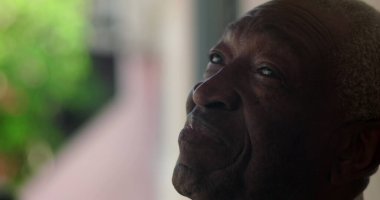 Afrika kökenli yaşlı bir adam düşünceli ve sakin bir tavırla yukarı bakıyor, hayatı yansıtıyor, arka planda yeşillik olan bir duvara yaslanıyor.