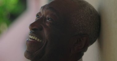 Afrika kökenli yaşlı bir adam, sıcak bir gülümseme, başını yaslanmış, bir duvara yaslanmış, açık havada canlı yeşillik, coşkulu bir sevinç, sıcaklık, ve bir karşılama davranışı ile