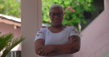 Afrikalı Latin kökenli bir kadın kısa beyaz saçlı ve gözlüklü, kendinden emin bir şekilde ayakta duruyor. Canlı doğal ortamda bireysellik ve sıcaklık yayıyor.