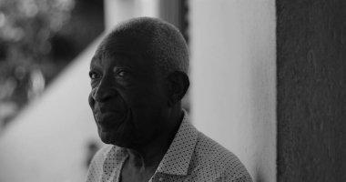 Düşünceli bir ifadeyle yan bakan yaşlı bir adam, açık havada bir duvara yaslanmış, bilgelik ve iç gözlem yayıyor, siyah ve beyaz