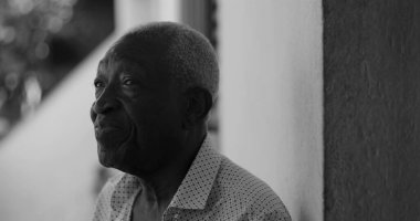 Düşünceli bir ifadeyle yan bakan yaşlı bir adam, açık havada bir duvara yaslanmış, bilgelik ve iç gözlem yayıyor, siyah ve beyaz