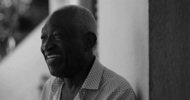 Afro-Amerikalı yaşlı bir adam duvara yaslanırken neşeyle gülümsüyor, mutluluk, memnuniyet ve sıcaklık yayıyor, siyah-beyaz.