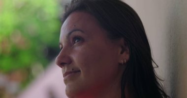 Huzurlu bir kadın zarif bir gülümsemeyle yukarı bakıyor, halka küpeler takıyor, bulanık yeşil arka plana yakın plan, özünde sakin ve sakin bir an yakalıyor.