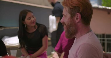 Bir grup arkadaş öğle yemeği için arka bahçede bir araya geldi, sakallı bir adamın yakın çekimi ile, arka planda gülümseyen bir kadın, neşe ve bağlantı iletiyor.