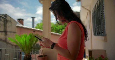 Dışarıda akıllı telefonu tutan, endişeli bir ifade sergileyen, düşüncelere dalıp alnına dokunan, arka planda bitkiler olan yerleşim yerlerinin yakınında duran bir kadın.