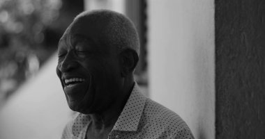 Siyah beyaz bir duvara yaslanmış, neşeyle gülen Afrikalı Amerikalı yaşlı bir adam, mutluluk, memnuniyet ve duygusal sıcaklık saçan, siyah beyaz bir şekilde yakalandı.