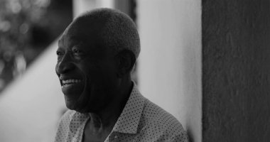 Afro-Amerikalı yaşlı bir adam duvara yaslanırken neşeyle gülümsüyor, mutluluk, memnuniyet ve sıcaklık yayıyor, siyah-beyaz.