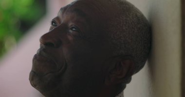 Afrika kökenli yaşlı bir adam, sıcak bir gülümseme, başını yaslanmış, bir duvara yaslanmış, açık havada canlı yeşillik, coşkulu bir sevinç, sıcaklık, ve bir karşılama davranışı ile