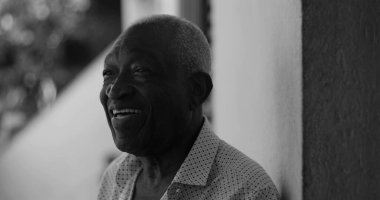 Afro-Amerikalı yaşlı bir adam duvara yaslanırken neşeyle gülümsüyor, mutluluk, memnuniyet ve sıcaklık yayıyor, siyah-beyaz.