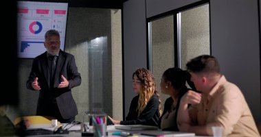 Kızgın işletme yöneticisi, meslektaşları ekranda gösterilen çizelgelerle kritik geri bildirimleri dinlerken toplantı odasında gergin bir toplantı sırasında ekibe işaret ediyor.