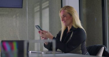 Modern ofis ortamında masa başında akıllı telefon kullanırken gülümseyen, iletişim kuran veya mesajları kontrol eden bir iş kadını