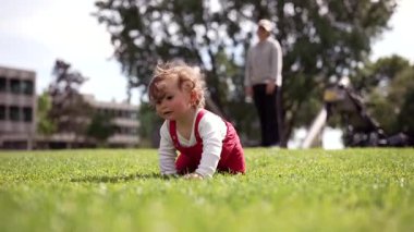 Çocuk parkta çimenlerde sürünüyor, yetişkin arka planda duruyor, merakla ve kararlılıkla çevreyi keşfediyor, erken bağımsızlık ve aile bağını simgeliyor.
