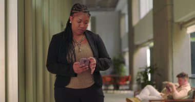 Ofis koridorunda yürürken akıllı telefon kullanan Afro-Amerikan kadın, arka grupta görünen oturma arkadaşları ile ekranda odaklanmış.