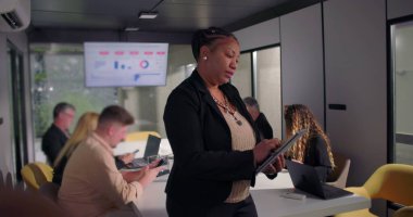 Dijital tablet kullanarak ofiste duran Afrikalı Amerikalı iş kadını, meslektaşları arka plandaki konferans masasında işbirliği yaparken işine odaklandı