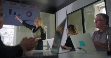 İş kadını ekranda el kol hareketi yaparken konferans odasında dizüstü bilgisayarları ve pencereleriyle meşgul profesyonellere sunum yapıyor.