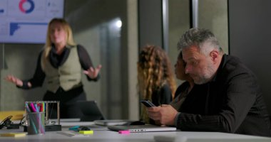 İş adamı ofis toplantısı sırasında gizlice telefonu kontrol ediyor. Kadın meslektaşları sunum yapıyor. Gündelik modern iş yeri dinamikleri ve çoklu görev ve sessiz takım çatışması.