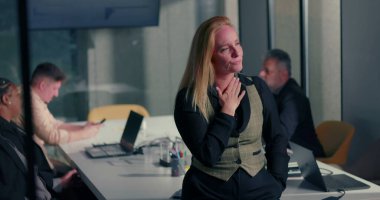 Blonde businesswoman with pensive and worried expression during office meeting, standing by conference table while colleagues work in background