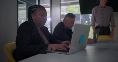 Kurumsal ofis toplantı odasındaki masada takım çalışması sırasında bilgisayarında meslektaşlarının yanında daktilo yazan Afrikalı Amerikalı iş kadını.