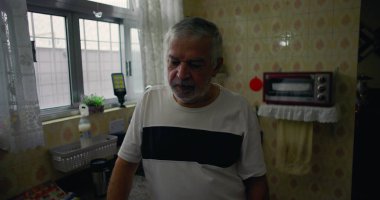 Mutfakta duran yaşlı bir adam, düşüncelere dalmış, evcil ortamda yalnızlığı ve sessizliği yansıtıyor.