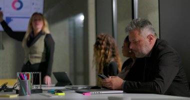 İş adamı ofis toplantısı sırasında gizlice telefonu kontrol ediyor. Kadın meslektaşları sunum yapıyor. Gündelik modern iş yeri dinamikleri ve çoklu görev ve sessiz takım çatışması.
