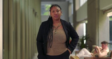 Parlak ofis koridorunda el ele tutuşan Afrikalı Amerikalı kadın, arka planda iş arkadaşları otururken doğrudan kameraya bakıyor.