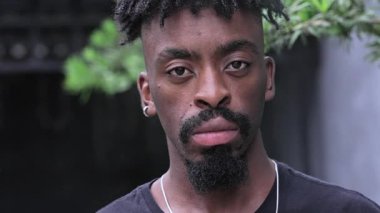 Sakallı siyah adam, kameraya bakan basit aksesuarlar takıyor. Sessiz ve tarafsız bir ifade kullanıyor.