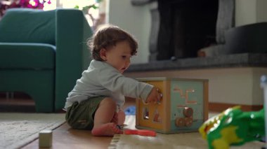 Yeni yürümeye başlayan çocuk yerde diz çöküyor. Aktivite küpüne tahta bloklar yerleştiriyor. Sakin aile ortamında merak ve neşeyle renkleri ve şekilleri keşfediyor.