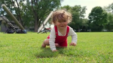 Şehir parkında çimenlerde sürünen çocuk, neşeli bir merakla dışarıyı keşfediyor, ağaçlar, oyun alanları ve yumuşak arka planda binalar, erken çocukluk özgürlüğünü simgeliyor.