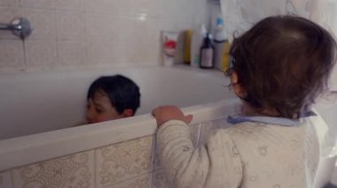 Meraklı bebek küvetin yanında durmuş, ablasının suda oynamasını izliyor. Aile hayatı çocukluk merakı, şefkat ve bağ kurma anı.