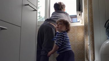 Daha yeni yürümeye başlayan çocuk, büyük kardeşinin kollarına sarılırken her ikisi de açık pencereden izleyip yakınlık, sıcaklık ve sessiz ortak merak üzerine kurulu nazik bir aile anı yaratıyor.