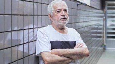 Uzaktan bakan yaşlı bir adam kolları çaprazlanmış bir şekilde duvara yaslanmış sessiz ve düşünceli bir şekilde duruyor.