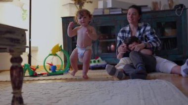 Küçük çocuk kameraya doğru sürünürken anne ve büyük kardeş arka planda dinleniyor, eğlenceli merakı yakalıyor ve gerçek aile hayatında bağımsızlığın ilk aşamalarını yakalıyor.