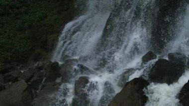 Büyük taşların arasından akan şelale, yüksek hızlı kamerayla çekilen Ultra yavaş çekimde yeşil yapraklarla çevrili.