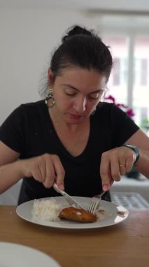 Kadın evde yemek masasında otururken tabakta yemek kesiyor odaklanmış ifade ve basit yemek ortamında ellere, çatal bıçaklara ve tabağa bakıyor.