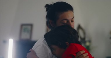 Bir anne, rahat bir anda çocuğunu kucağına alır. Çocuk, onun omzunda huzur içinde dinlenir. Onların kucaklaşması, sıcaklıkla ve güvenle dolu hassas, samimi bir sahne yaratır..