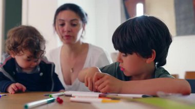 Anne, küçük çocuk ona yaslanıp yaratıcılık, yakınlık ve ortak ilgi odaklı nazik ve sakin bir aile anı oluştururken, büyük çocuğunun çizim üzerinde yoğunlaştığını gözlemler.