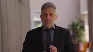 Orta yaşlı bir adam kravatını düzeltiyor sonra kameraya bakıyor, sakin ve içsel bir jest hazırlığı, özgüveni ve sessiz farkındalığı simgeliyor.