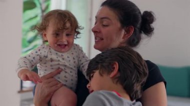 Anne, yeni yürümeye başlayan çocuğu ve büyük çocuğu sakin bir ifadeyle kucaklarken, yeni yürümeye başlayan çocuk merakla ağza dokunuyor ve yumuşak bir duygusal sıcaklık ve güven anı yaratıyor.