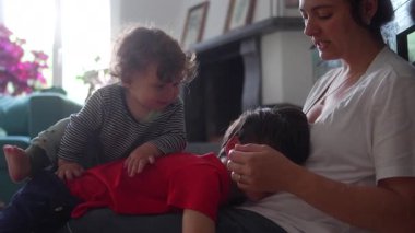 Küçük bir çocuk, annesinin kucağında yatan büyük bir kardeşin üzerine çıkıyor. Sahne, yumuşak ışıklı oturma odasında neşeli aile yakınlığının sıcak ve samimi bir anını yakalıyor.. 