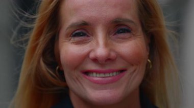 Orta yaşlı kadının nazikçe gülümsemesi, sıcaklığın, özgüvenin ve açık havada duygusal açıklığın ifadesi.