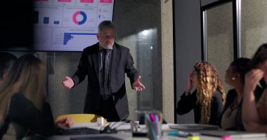 Kızgın işletme yöneticisi, meslektaşları ekranda gösterilen çizelgelerle kritik geri bildirimleri dinlerken toplantı odasında gergin bir toplantı sırasında ekibe işaret ediyor.