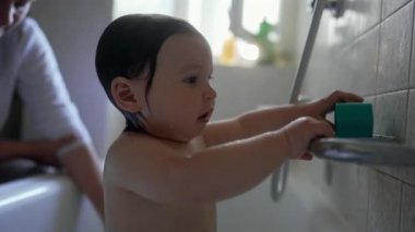 Bebek küveti keşfederken dikkatli bir konsantrasyonla su başından aşağı akarken oyun, duyusal öğrenme ve nazik aile bakımıyla dolu sıcak ve dalgın bir an yaratıyor.