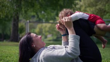 Anne çimenlerin üzerinde uzanıyor ve kırmızı tulumlu bebekle neşeyle oynuyor. Güneşli bir günde huzurlu yeşil parkta birlikte gülüyor ve neşeli anları paylaşıyor.