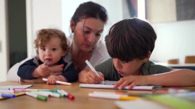 Anne, büyük çocuk çizime odaklanırken iki çocuğu da izliyor ve küçük çocuk da işaretleri inceleyerek, ortak yaratıcılık ve nazik aile bağları için sakin bir ev yaratma anı oluşturuyor.