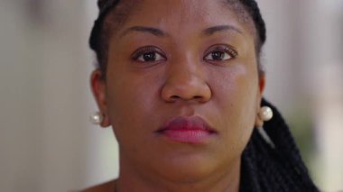 Afro-Amerikan kadın portresi, sakin bir ifadeyle doğrudan kameraya bakıyor, doğal ışık yüzüne vurgu yapıyor ve saçları yumuşak bulanık arka planda örüyor.