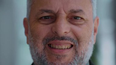 Gri sakallı, yorgun orta yaşlı bir adamın yakın portresi tedirgin bir gülümseme sergiliyor, yüzeyin altında huzursuzluk ve duygusal gerginlik gösteriyor ve neşeli görünmeye çalışıyor.