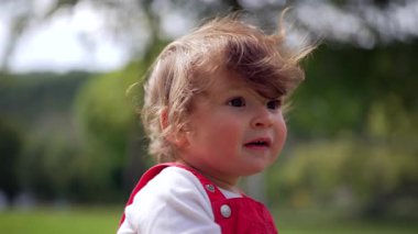 Kırmızı tulumlu bebek, saçı rüzgarlı, tuhaf bir şekilde mesafeye bakıyor, parkta doğal yakın plan portresi, masumiyet, merak ve çocukluk duygularını aktarıyor.