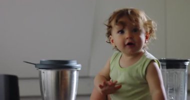 Bir çocuk mutfak tezgahının üzerinde meraklı gözlerle ve neşeli bir ifadeyle sürünerek ilerler. Evdeki aktif ve keşifsel bir anda kameraya doğru eğilir.