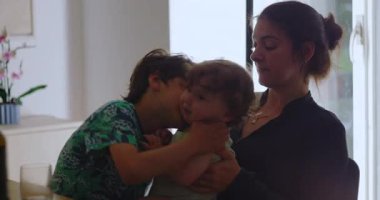 Anne yeni yürümeye başlayan çocuğu rahatlatırken, büyük çocuk da oyuncu etkileşimlere katılıyor, aile içinde hassas duygusal yakınlık ve ev ortamında sıcaklık yaratıyor.