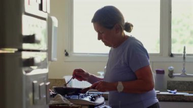 Evde yemek pişiren yaşlı bir kadın, pencerenin yanındaki tavada yemek karıştıran, günlük rutin hareketler yapan ve parlak ev mutfağında yoğunlaşan bir kadın.